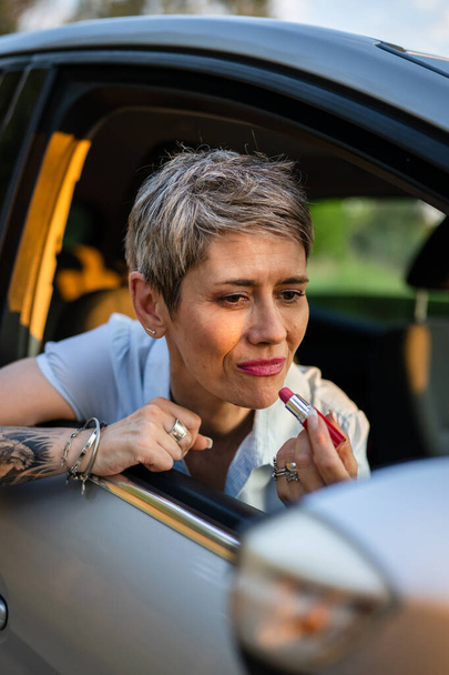 Eine reife kaukasische Geschäftsfrau sitzt im Auto und setzt Lippenstift, der ihr Gesicht repariert, während sie im Sommertagabend wartet, echte Menschen kopieren graue Haare - Foto, Bild