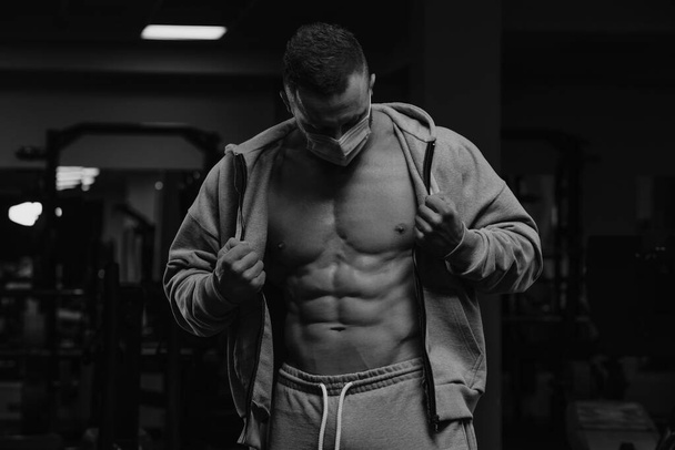 A bodybuilder in a face mask to avoid the spread of coronavirus is opening his zipped hoodie to demonstrate his athletic physique. A sporty guy in a surgical mask is posing after a workout in a gym. - Photo, Image