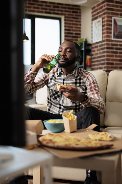 Веселый мужчина пьет алкогольное пиво из бутылки и ест еду на вынос из пакета быстрого питания, смотрит кино по телевизору. Наслаждаясь напитками и едой на вынос перед телевизором. - Фото, изображение