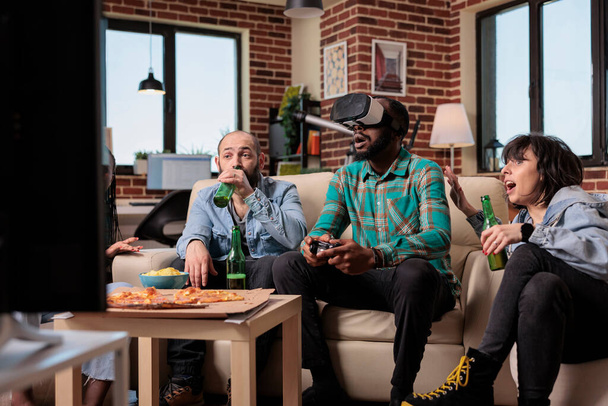 Multiethnic group of friends using virtual reality glasses to play video games on television. Having fun with beer bottles and snacks at house oarty, playing competition with vr goggles. - 写真・画像