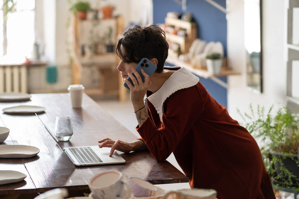 Молода жінка власник гончарного бізнесу використовує ноутбук і розмовляє з клієнтом по телефону під час роботи в майстерні. Жінка-художниця заробляє гроші на продажу кераміки. Менеджер керамічного магазину перевіряє онлайн замовлення
 - Фото, зображення