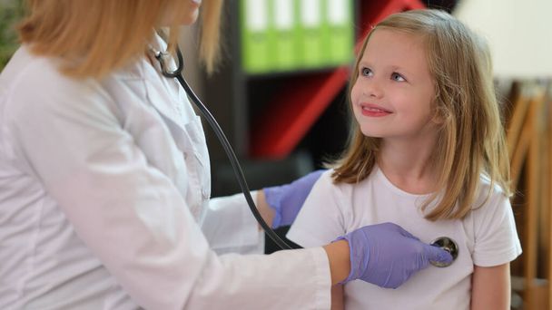 Portrait of smiling girl at doctor appointment in clinic office. Woman pediatrician examining little child with stethoscope. Healthcare, pediatrics and medicine concept - Фото, изображение