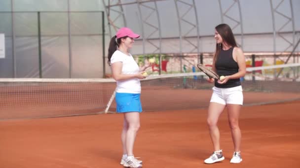 Two women stand on a tennis court - playing with balls and rackets and talking. Mid shot - Кадры, видео