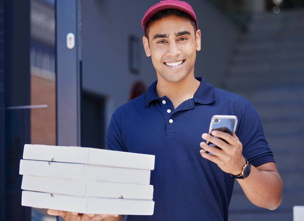 Ordering online has never been easier. Portrait of a young delivery man using a cellphone while delivering pizzas - Фото, изображение