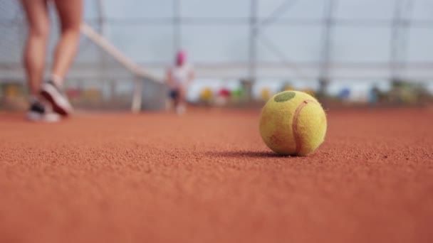 Tennis ball on the ground and two women tennis players warming up on the background. Mid shot - Filmagem, Vídeo