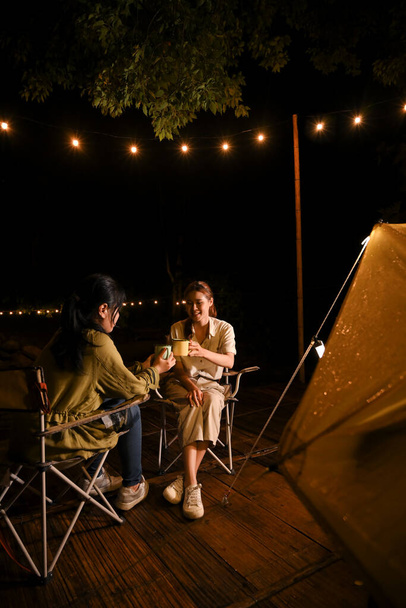 Two millennial Asian female best friends talking, sipping hot coffee and enjoy camping together at the campground at night. - Foto, imagen