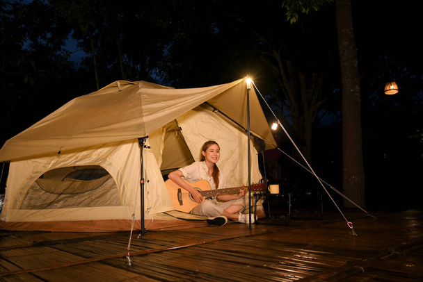 Mulher asiática jovem atraente tocando guitarra na frente da tenda teepee no acampamento à noite. Conceito de atividade exterior. - Foto, Imagem