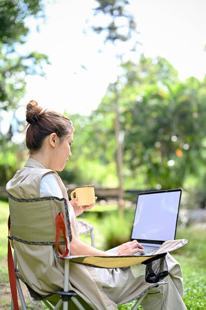 Atraktivní tisíciletá asijka sedící na tábořišti, popíjející kávu a používající notebook k práci na svém úkolu. Koncept kempování outdoorové aktivity - Fotografie, Obrázek