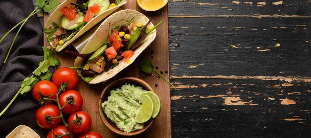 Tacos, tomatoes and tasty guacamole on black wooden background, top view - Фото, изображение