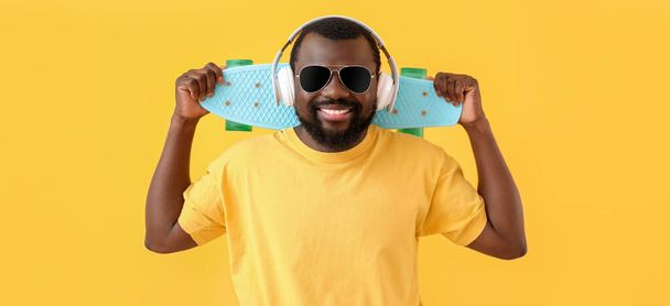 Schöner afroamerikanischer Mann mit Kopfhörer und Skateboard auf gelbem Hintergrund - Foto, Bild
