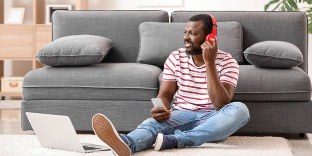 Handsome African-American man with phone and laptop listening to music at home - Photo, image