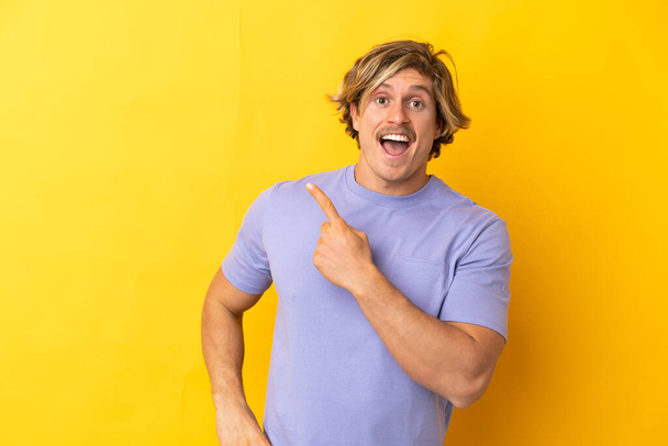 Handsome blonde man isolated on yellow background surprised and pointing side - 写真・画像