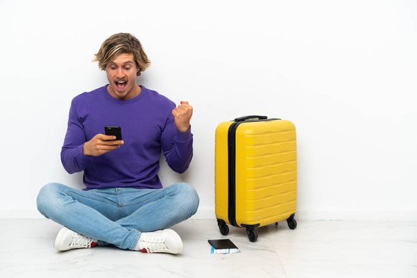 Молодой блондин с чемоданом, сидящий на полу удивлен и отправляет сообщение - Фото, изображение