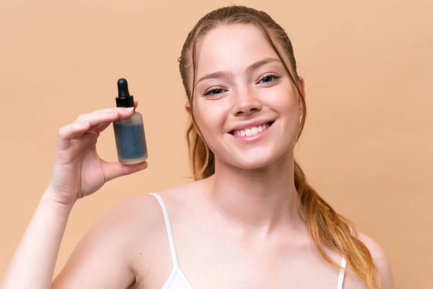 Young caucasian girl isolated on beige background holding a serum while smiling. Close up portrait - Foto, Imagem