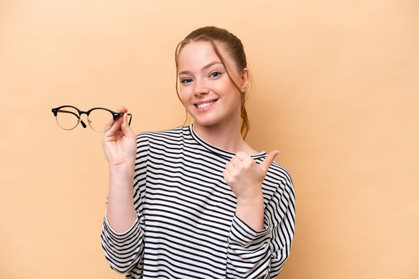 Bej arka planda izole edilmiş gözlüklü beyaz kız bir ürünü sunmak için yan tarafı işaret ediyor. - Fotoğraf, Görsel