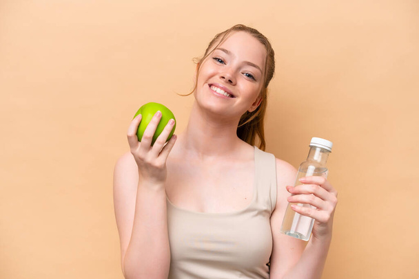 Young caucasian girl isolated on beige background with an apple and with a bottle of water - Foto, imagen