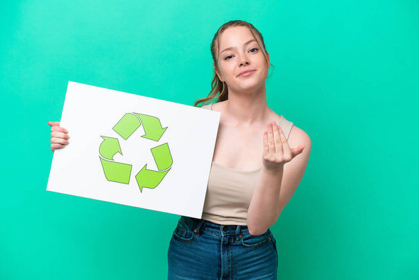Young caucasian woman isolated on green background holding a placard with recycle icon and doing coming gesture - Foto, imagen
