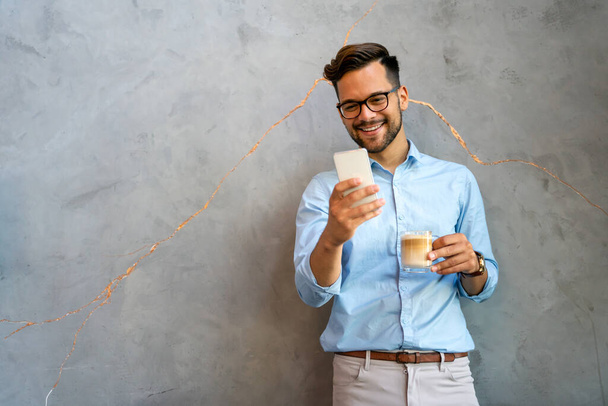Portrait of a smiling young handsome business man using smartphone and holding cup of coffee - Photo, Image