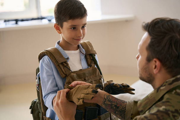 Child looking at his father while he zipping up glove on his hand during military gear training - Photo, image