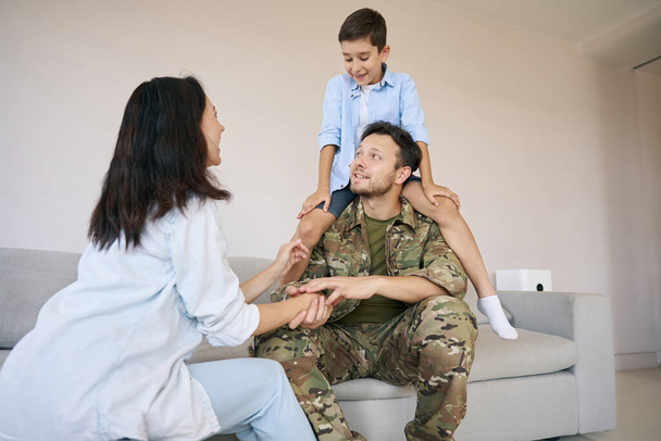 Man returning from military service sitting on couch with kid on his neck while meeting with his wife - Photo, Image