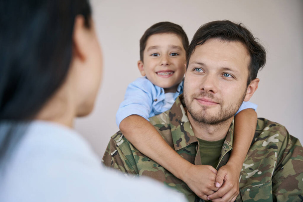 Filho abraçando o pescoço soldado por trás e sorrindo para a câmera, enquanto o homem olhando para sua esposa - Foto, Imagem