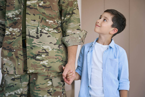 Proud boy looking up at his soldier father while holding his hand at home - Foto, afbeelding