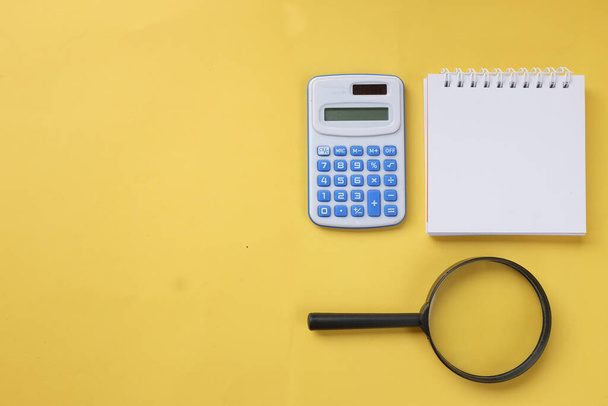 copy space white spiral notepad and magnifying glass ,blue Calculator,pencil on a yellow background - Foto, imagen