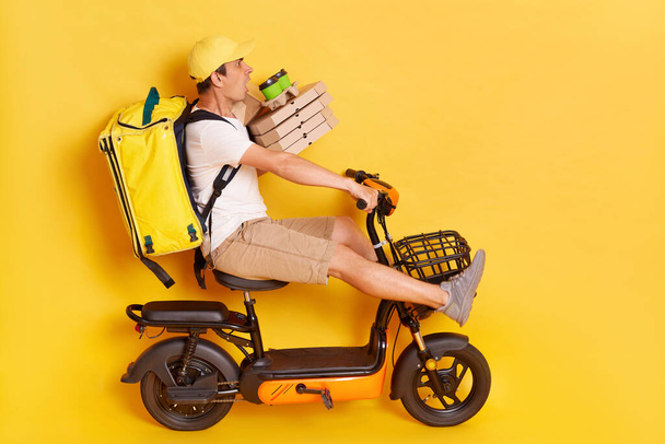 Side view of shocked scared man wearing white t shirt and cap riding electric scooter with backpack, holding pizza boxes and coffee in disposable cups, road accident, isolated over yellow background. - Photo, Image