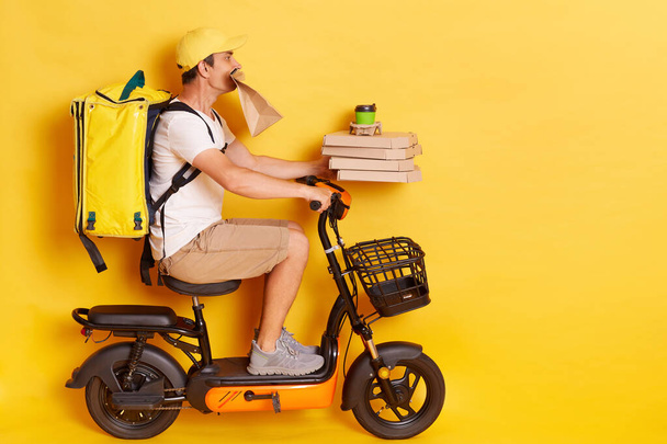Profile portrait of young man with backpack wearing white t shirt riding e-scooter with pizza boxes and coffee, holding paper parcel in teeth, delivering lots orders, isolated over yellow background. - Foto, Bild