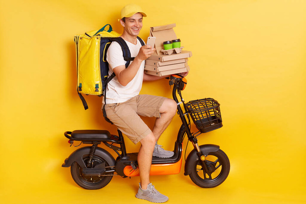 Young courier in white t shirt, pizza delivery man with thermo backpack and phone on moped isolated on yellow background. Fast transport express, home food delivery. Online order. - Фото, зображення