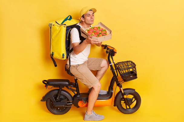 Mmm, how delicious. Satisfied delivery man enjoys pleasant smell from freshly baked pizza on cardboard box, has good appetite, transports delicious snack for clients, poses over yellow background. - Photo, image