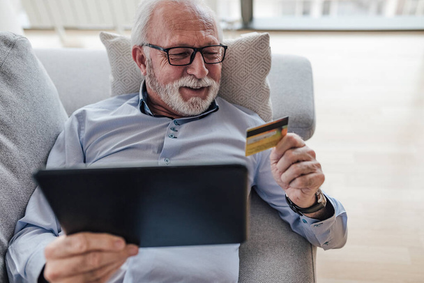 Smiling senior professional looking at credit card. Elderly manager making online payment through digital tablet. He is lying on sofa at home office. - Φωτογραφία, εικόνα