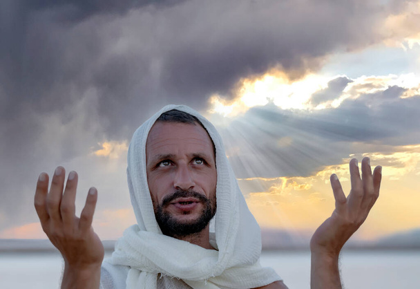 Caucasian man wearing a headscarf raise his hands up to sunset sky and looking up. Christian worship, God, Jesus face, religion or freedom concept. Bearded arab guy with green eyes prayers with natural face. Man Raising His Hands to Heaven - 写真・画像