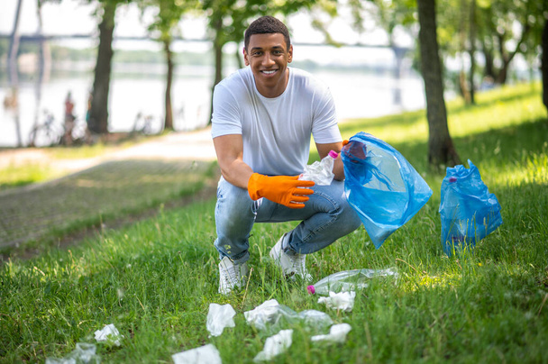 Useful activity. Young dark-skinned man smiling at camera crouching picking up waste from grass putting trash in bag - Φωτογραφία, εικόνα