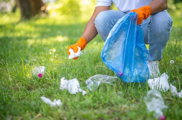 Garbage collection. Man in gloves crouched collecting garbage from grass into bag in park on sunny day no face - Foto, Imagen