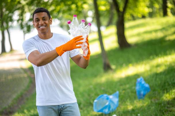 Save planet. Dark-skinned guy with toothy smile pointing at camera plastic garbage standing in green park - 写真・画像
