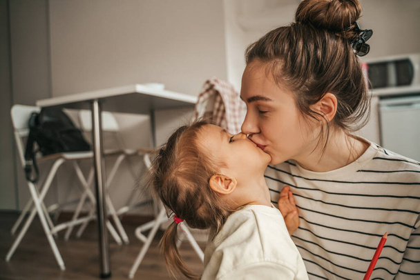Loving beautiful young female parent giving a tender kiss to her child in the kitchen - Фото, зображення
