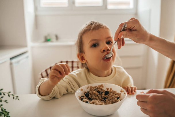 Caring parent giving a spoon of oatmeal to a little girl seated at breakfast at the kitchen table - 写真・画像