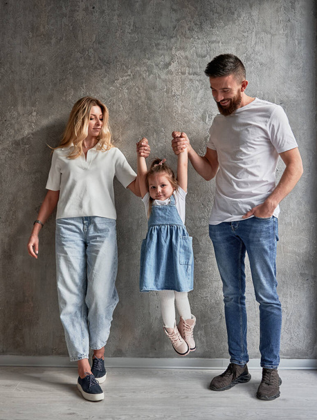 Full length of happy family with child standing against the wall in newly built apartment. Woman and man holding hands of adorable daughter and lifting kid in the air in new home. - Φωτογραφία, εικόνα
