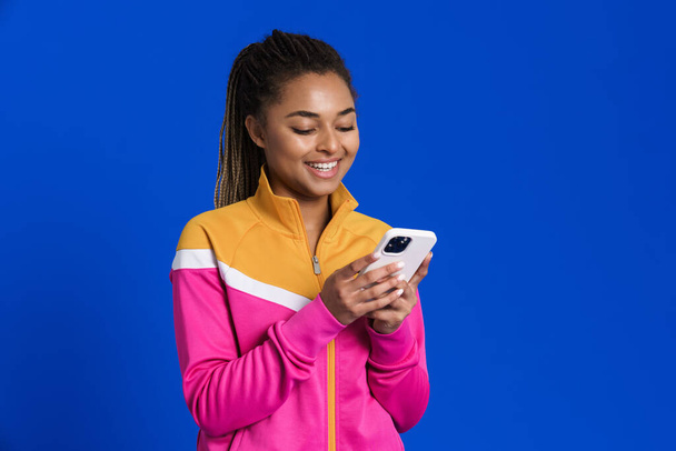 Black young woman laughing while using mobile phone isolated over blue background - Φωτογραφία, εικόνα