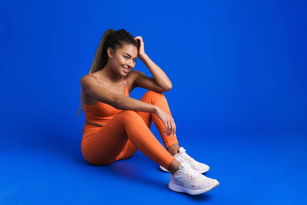 Black young sportswoman smiling while sitting on floor isolated over blue background - Φωτογραφία, εικόνα
