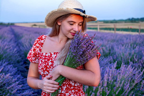 A beautiful blonde woman with her eyes closed is smelling a freshly picked bouquet of lavender flowers, in a picturesque lavender field. Aromatherapy, relaxing. - 写真・画像