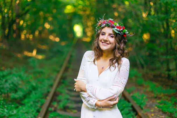 beautiful girl in a white dress with a wreath on her head embraces herself and smiles. railway and green trees - Foto, imagen