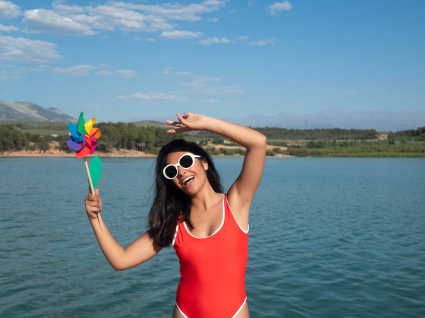 Delighted female in swimsuit holding pinwheel while standing on shore near rippling sea on summer day in coastal area - 写真・画像