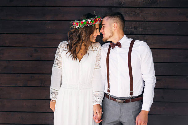 couple near the wooden wall hold hands, closed their eyes and smile. couple in festive clothes. girl with a wreath on the head - Foto, Bild