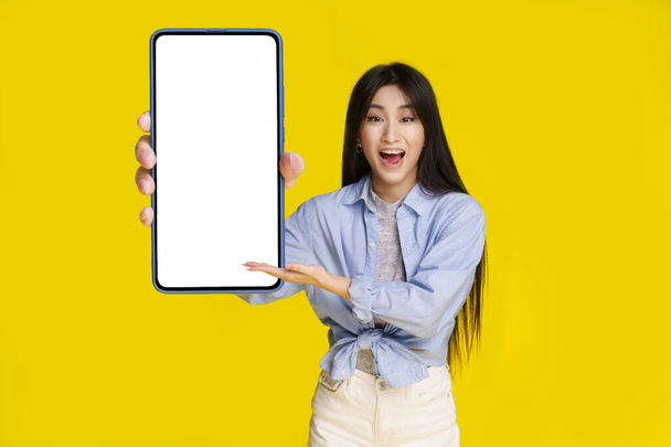 Big smartphone with white screen in hand of beautiful asian girl happy to introduce new app, game, win, isolated on yellow background. Mobile app advertisement product placement.  - Photo, Image