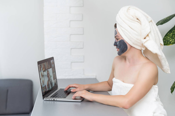 Nuori nainen pyyhe päässä ja musta kasvojen savi naamio istuu kotona ja käyttää kannettavaa tietokonetta. Kotona oleskelu, karanteeni, etäopetus, verkko-opetus, teknologia, moderni elämäntapa. - Valokuva, kuva