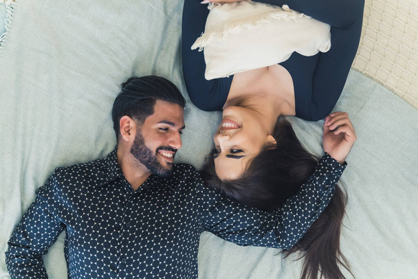 Smiling couple having fun while laying on bed with white sheets and looking at each other. Overhead view.  - Photo, Image