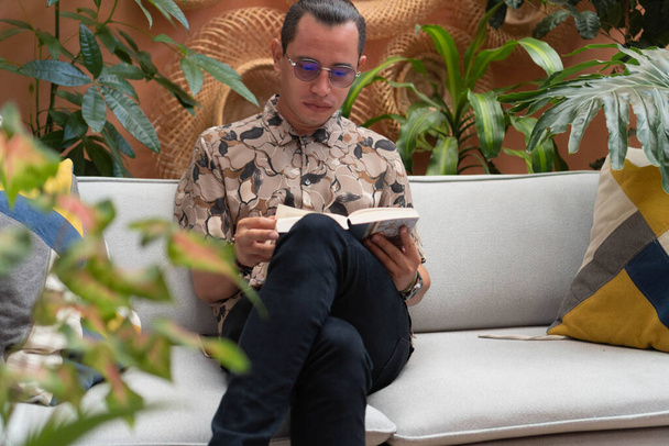 Latin férfi ül egy karosszékben és könyvet olvas. A fiatalember könyvet olvas a kényelmes lakásában.. - Fotó, kép