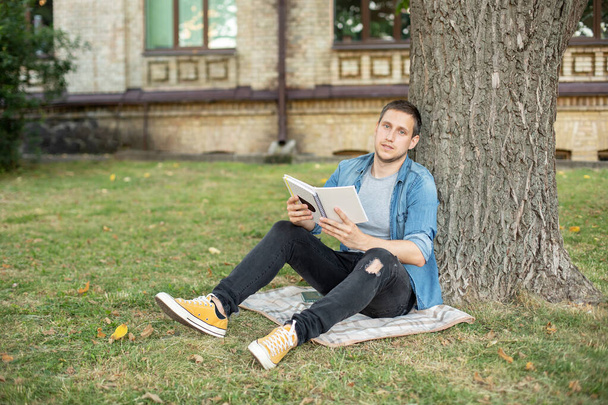 Молодой человек расслабляется и читает книгу, сидя на газоне. Счастливчик сидит на зеленой траве и учится дистанционно. Студент-взрослый читает в парке. Наслаждаюсь студенческой жизнью. улыбающийся человек, работающий на открытом воздухе  - Фото, изображение